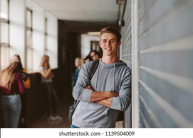 Portrait Of Handsome Young University Student Standing And Leaning To A Wall In College Corridor With Other Students At The Back. Caucasian Male Student In High School Campus.