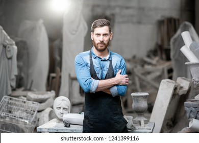Portrait of a handsomme sculptor in blue t-shirt and apron standing in the studio with old sculptures on the background