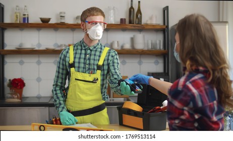 Portrait Of Handyman And Woman Customer In Protective Masks In Kitchen. Plumber Holding Terminal For Client To Pay With Credit Card For Repair Services