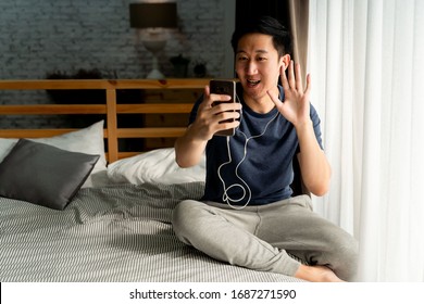 Portrait of happy 30s aged Asian man in casual clothing making facetime video calling with smartphone at home. He's waving at people on phone screen. Social distancing concept