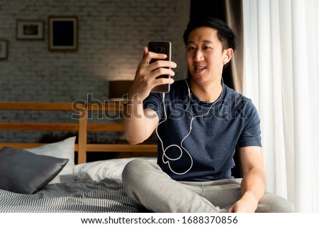 Portrait of happy 30s aged Asian man in casual clothing making facetime video calling with smartphone at home. He's waving at people on phone screen. Using conferencing meeting online app concept