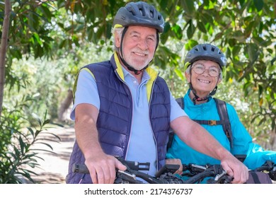 Portrait Of Happy Active Caucasian Senior Couple With Electric Bicycles Running Outdoors In The Park. Two Smiling Seniors Wearing Cycling Helmet Enjoying A Healthy Lifestyle In Nature