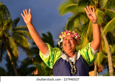 Portrait Of Happy Active Senior Polynesian Pacific Islander Woman On Tropical Beach With Palm Trees In The Background Waving With Her Hands Welcome Hello. Real People. Copy Space