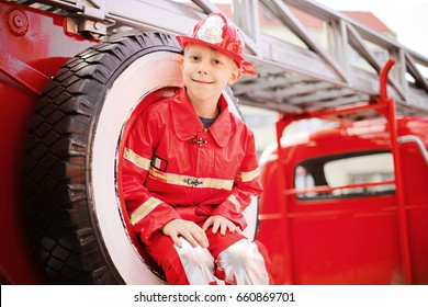 Portrait Of Happy Adorable Child Boy With Fireman Hat Playing Outside Siting In Spare Wheel Of Old Shiny Vintage Red Fire Truck. Dreaming Of Future Profession. Fire Safety, Life Protection Lessons