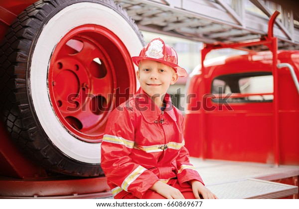 Portrait of Happy Adorable Child Boy with Fireman
Hat Playing Outside siting near spare wheel of old shiny vintage
red fire truck. Dreaming of future profession. Fire safety, Life
Protection lessons