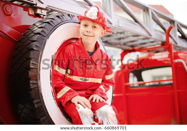 Portrait of Happy Adorable Child Boy with Fireman
Hat Playing Outside siting in spare wheel of old shiny vintage red
fire truck. Dreaming of future profession. Fire safety, Life
Protection lessons