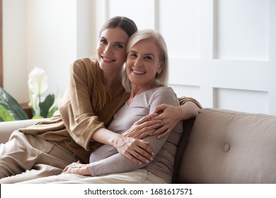 Portrait Happy Adult Daughter And Older Mother Hugging And Holding Hands, Sitting On Couch At Home, Young Woman And Mature Mum Or Grandmother Looking At Camera, Two Generations Family Photo