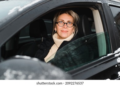 Portrait Of Happy Adult Woman Driving Car In Winter Season, Outdoors. Woman Driver Inside Black SUV Through Window Looking At Camera.