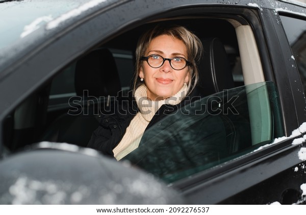 Portrait of happy adult woman driving car in\
winter season, outdoors. Woman driver inside black SUV through\
window looking at\
camera.