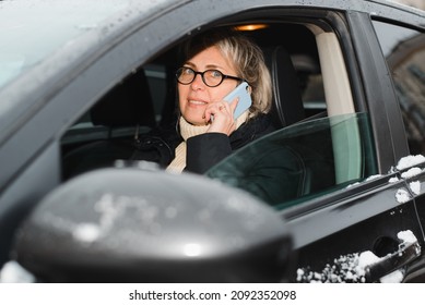 Portrait Of Happy Adult Woman Talking On Cell Phone While Driving Car In Winter Season, Outdoors. Smiling Female Driver With Smartphone Sitting Inside Black SUV And Looking At Camera.