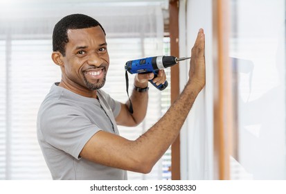Portrait of happy African American black man using electric drill on white wall. DIY, do it yourself hobby, handyman worker using drill while working on construction site. Home interior builder moving