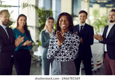 Portrait of a happy African American businesswoman celebrating success and applauding, surrounded by colleagues in the office. Moment of an achievement and camaraderie among the group. - Powered by Shutterstock