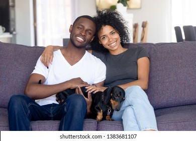 Portrait Of Happy African American Couple Hug Sitting On Couch With Cute Dog, Smiling Black Husband And Wife Pose With Dackel, Relaxing Together On Sofa, Young Family With Pet Rest In Own Living Room