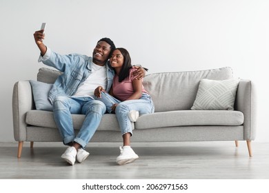 Portrait of happy African American couple taking selfie together at home indoors. Excited man and woman posing and holding smartphone, sitting on the couch in living room, free copy space - Powered by Shutterstock