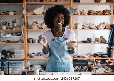 Portrait of a happy african american craftswoman standing at pottery workshop in studio with earthenware in hands and smiling at the camera. Multicultural pottery class attendee having wine on class. - Powered by Shutterstock