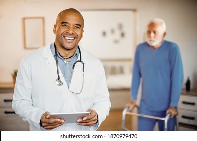 Portrait Of Happy African American Doctor Using Digital Tablet And Nursing Home And Looking At Camera. Senior Patient Is In The Background. 