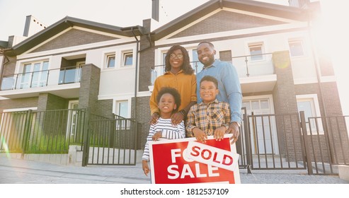 Portrait Of Happy African American Family With Small Children Standing At New House At Suburb And Smiling With Sold Table In Hands. 