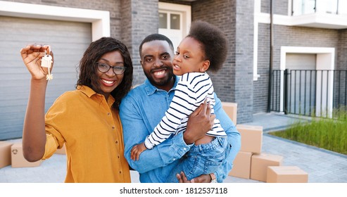 Portrait Of Happy African American Family With Small Cute Girl On Hands Standing At Big House On Suburb, Smiling And Showing Key. 