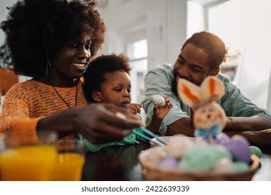 Portrait of a happy african american family of three painting Easter eggs together while sitting at wooden table in cozy home interior and enjoying DIY art. Copy space. - Powered by Shutterstock