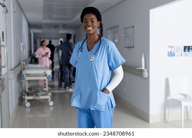 Portrait of happy african american female doctor wearing scrubs in corridor at hospital. Hospital, medicine, healthcare and work, unaltered. - Powered by Shutterstock