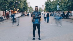 Portrait Of Happy African American Guy Wearing Stylish Jeans And Leather Jacket Standing Alone In Street Downtown, Smiling And Looking At Camera With Crowd Moving By.