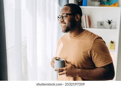 Portrait of happy African American guy holding cup of coffee looking out the window at home - Powered by Shutterstock