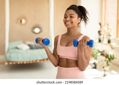 Portrait Of Happy African American Lady Doing Dumbbell Workout At Home, Working On Arms Strength And Smiling, Copy Space. Fit Woman Lifting Blue Fitness Dumbells Up Over Bedroom Interior