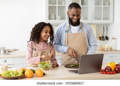 Portrait of happy African American man and little girl cooking in the kitchen and watching food video blog on laptop or reading recipe online using computer, cutting vegetables, preparing dinner