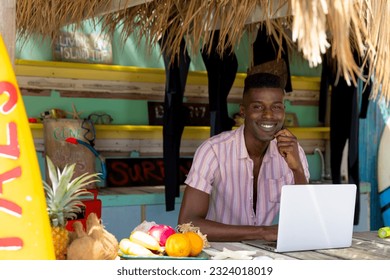 Portrait of happy african american man using laptop behind counter of surf hire beach shack. Local business, business owner, communication, hobbies, sport, surfing, summer and vacation, unaltered. - Powered by Shutterstock