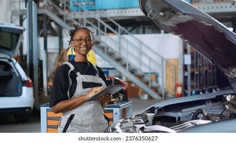 Portrait of happy african american mechanic doing car maintenance in repair shop using tablet to keep track of operations. Expert in garage looking over checklist on device while fixing vehicle - Powered by Shutterstock