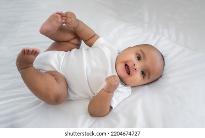 Portrait Of Happy African American Mixed Race Cute Girl Baby Lying Down On White Bed And Look On Camera In A Bedroom At Home. Top View Of Little Newborn Infant Smiling Laughing And Lying On Duvet.