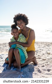 Portrait Of Happy African American Mother And Daughter Sitting On Sand At Beach During Sunny Day. Unaltered, Family, Lifestyle, Togetherness, Enjoyment And Holiday Concept.