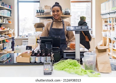 Portrait Of Happy African American Saleswoman Or Small Business Owner Wearing Apron In Sustainable Small Local Business. Young Afro Woman At Cashdesk With Scales In Local Zero Waste Grocery Store