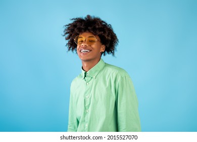 Portrait Of Happy African American Teen Guy In Mint Shirt And Yellow Sunglasses Posing On Blue Studio Background. Enjoying Summertime Vacation And Smiling To Camera