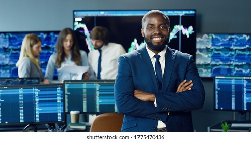 Portrait Of Happy African American Trader Or Broker Working At Stock Exchange Office On Background Of His Business Team. Investment Entrepreneur Trading Concept