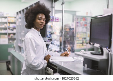 Portrait Of A Happy African American Woman Pharmacist Writing Prescription At Workplace In Modern Pharmacy, Looking At Camera With Smile