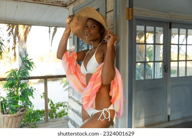 Portrait of happy african american woman in bikini and sunhat standing on porch of beach house. Summer, fashion and beauty, free time and vacations, unaltered. - Powered by Shutterstock