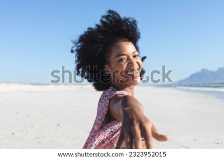 Portrait of happy african american woman smiling with hand outstretched on sunny beach. Summer, wellbeing, freedom, relaxation and vacation, unaltered.