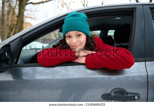 Portrait of a happy African American woman\
travelling by car, leaning on the driver door with opened windows\
and cutely smiles looking at\
camera.