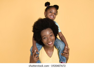 Retrato de una feliz bebé africana sentada en los hombros de su madre y divirtiéndose, riéndose ante la cámara en un aislado fondo amarillo Foto de stock