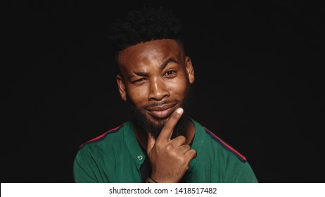 Portrait of happy african male looking at camera with a raised eyebrow. African man thinking with fingers on chin isolated on black background.