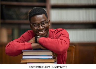 Portrait of happy african student wearing spectacles sitting in library leaning on pile of books. Smiling black young man at college library. Happy american guy at university with copy space.