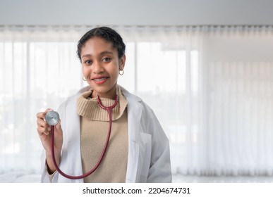 Portrait Of Happy African Woman Professional Doctor In Uniform Standing Holding Stethoscope And Look At Camera In White Room At Hospital. Black Female Medical Wear Braces On Teeth And Smile In Clinic.