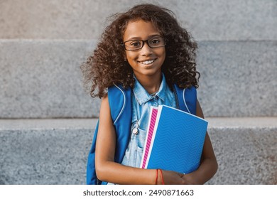 Portrait of happy african young preteen girl elementary middle school pupil in eyeglasses with backpack holding notebook books outside the primary school. Education concept - Powered by Shutterstock