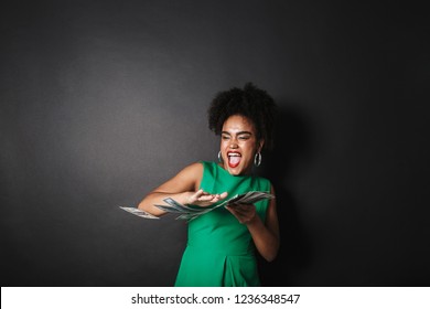 Portrait of a happy afro american woman wearing dress standing over black wall, throwing paper money away
