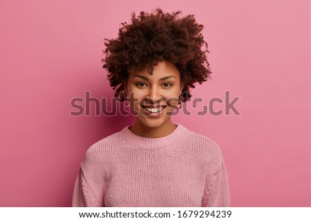 Portrait of happy Afro American woman has curly hairstyle, smiles and pleased expression, wears casual pink jumper in one tone with background, expresses happiness, glad to meet with close friend