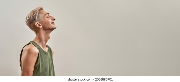 Portrait of happy androgynous transgender young man lifting head and smiling with eyes closed while posing isolated over light background. Pride, lgbtq, generation Z concept. Side view