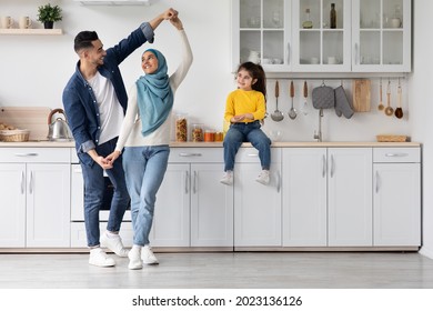 Portrait Of Happy Arab Family Of Three Having Fun In Kitchen Interior, Cute Smiling Little Middle Eastern Girl Sitting On Table And Watching Her Parents Dancing, Enjoying Time At Home Together