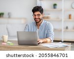 Portrait of happy arab freelancer man sitting at desk with laptop computer at home office, looking and smiling at screen, copy space. Young entrepreneur guy enjoying remote work