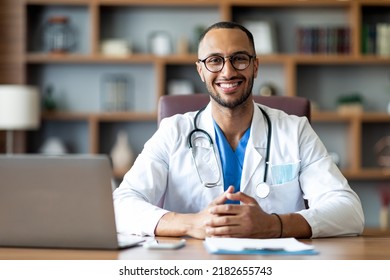 Portrait of happy arabic doctor handsome young man in workwear posing at modern clinic, sitting at workdesk with laptop and medical chart, cheerfully smiling at camera, copy space - Powered by Shutterstock